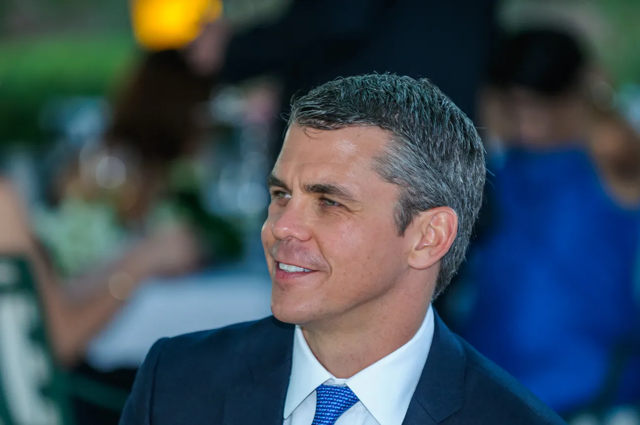 Smiling man in a dark suit and blue tie looking to the side with a blurred background of people.