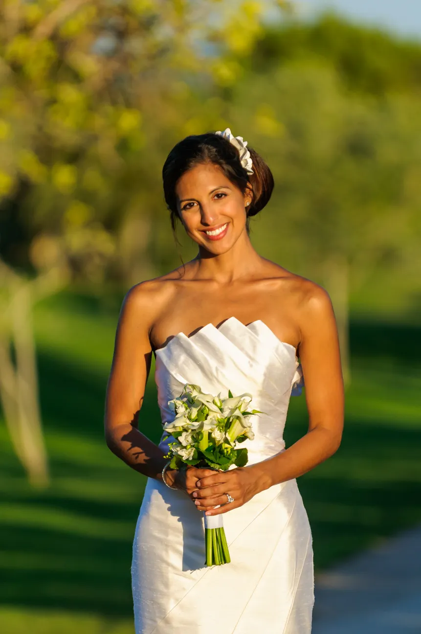 Smiling bride in a strapless white wedding dress holding a bouquet of white flowers outdoors.