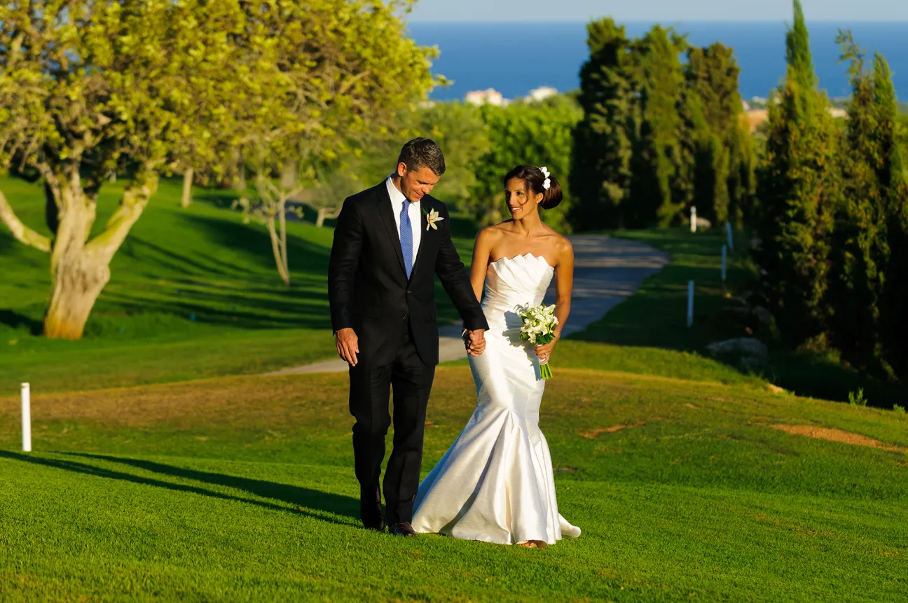 Bride in white gown holding bouquet and groom in black suit walking hand in hand on a grassy path with trees and ocean in the background.