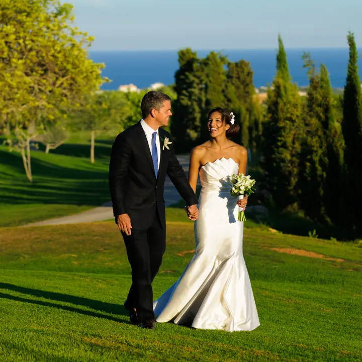 Bride and groom holding hands and walking on green grass with trees and ocean in the background during sunset.