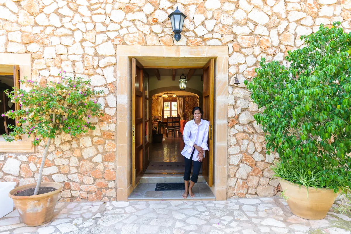 Smiling woman standing barefoot in the open doorway of a stone house with potted plants on each side.