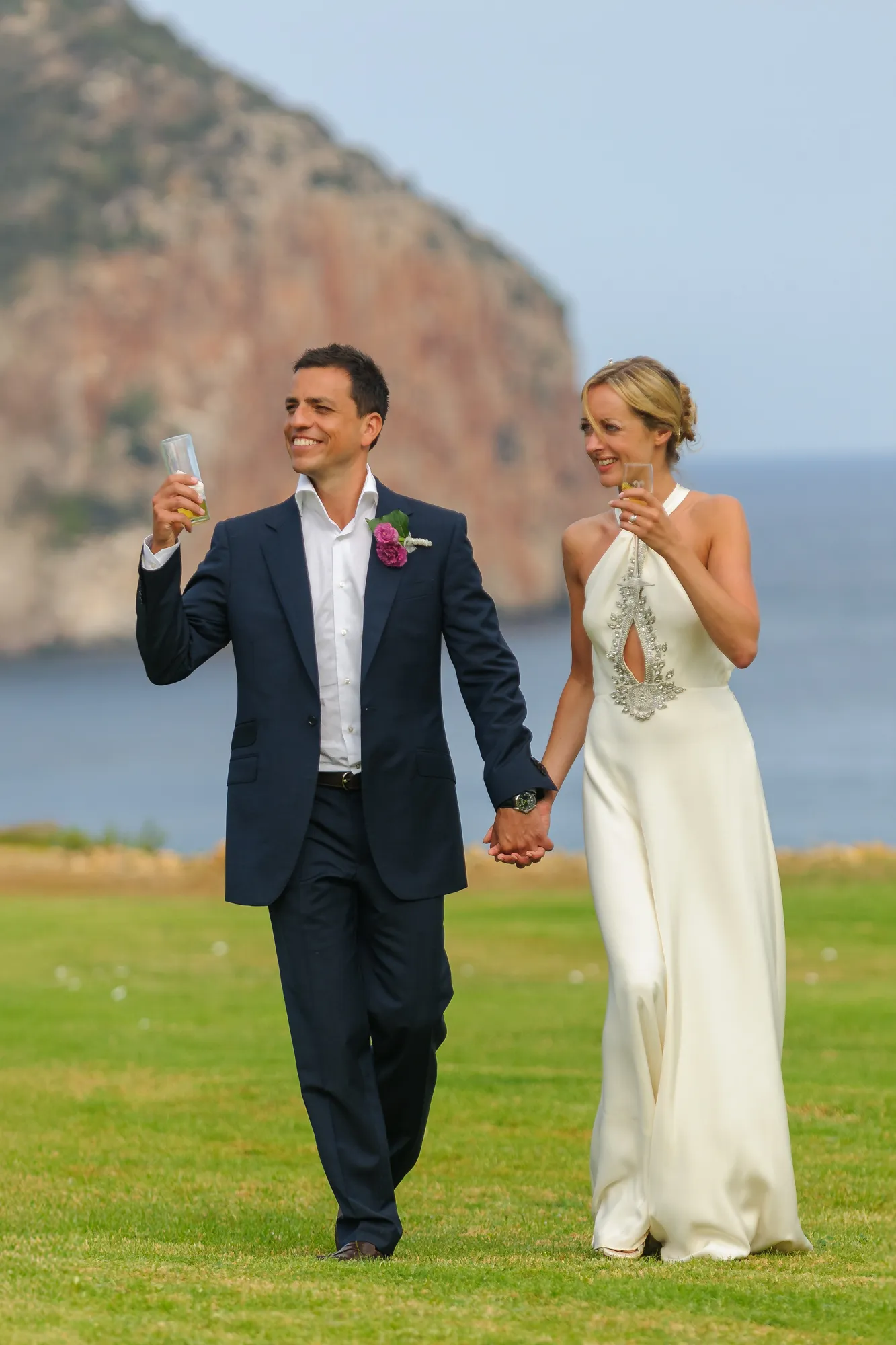 Smiling bride and groom holding hands and glasses of drink, walking on green grass with a rocky shoreline and ocean in the background.