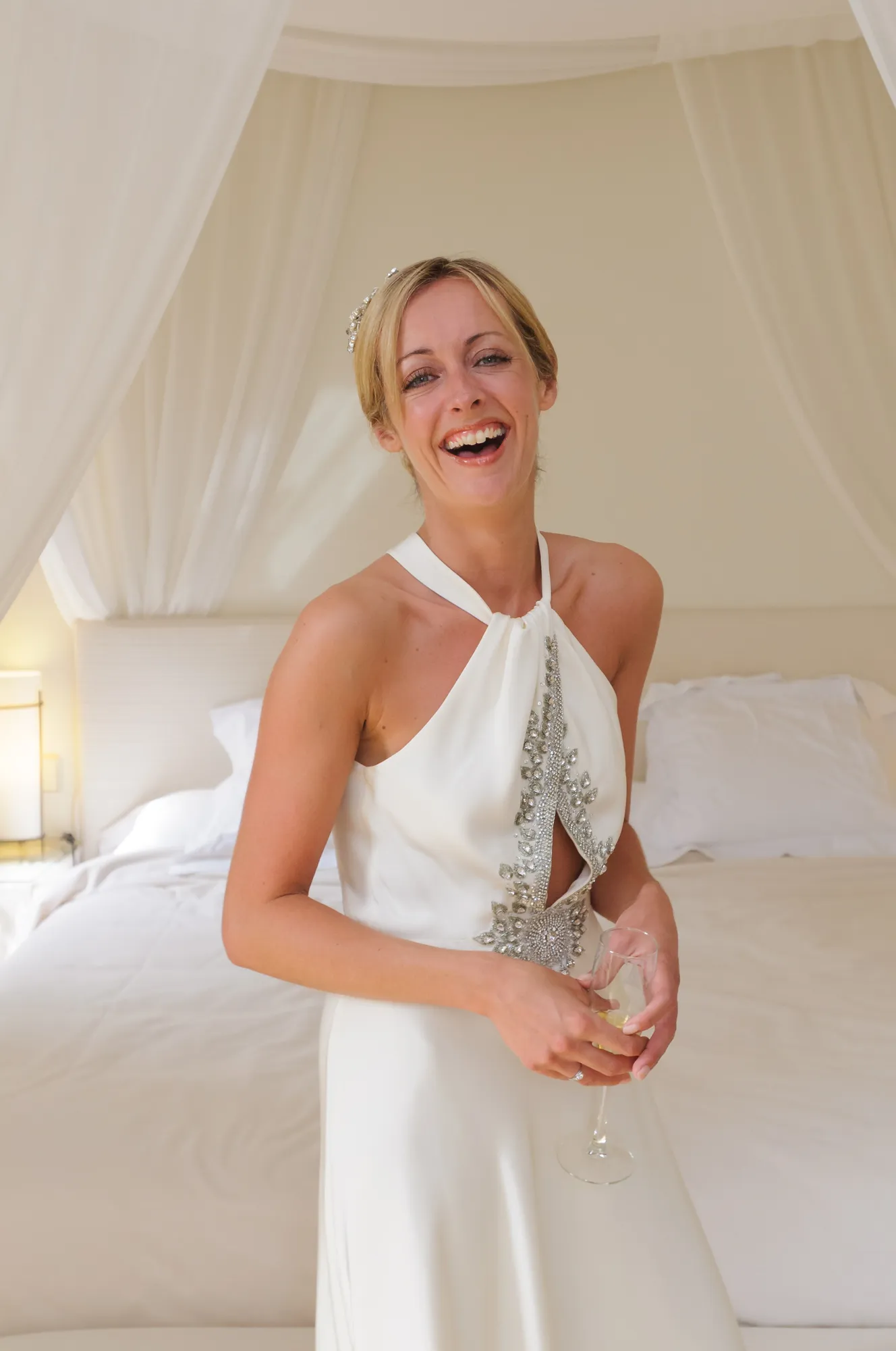 Smiling bride in a white halter wedding dress with beaded embellishments holding a glass of champagne in a softly lit bedroom.