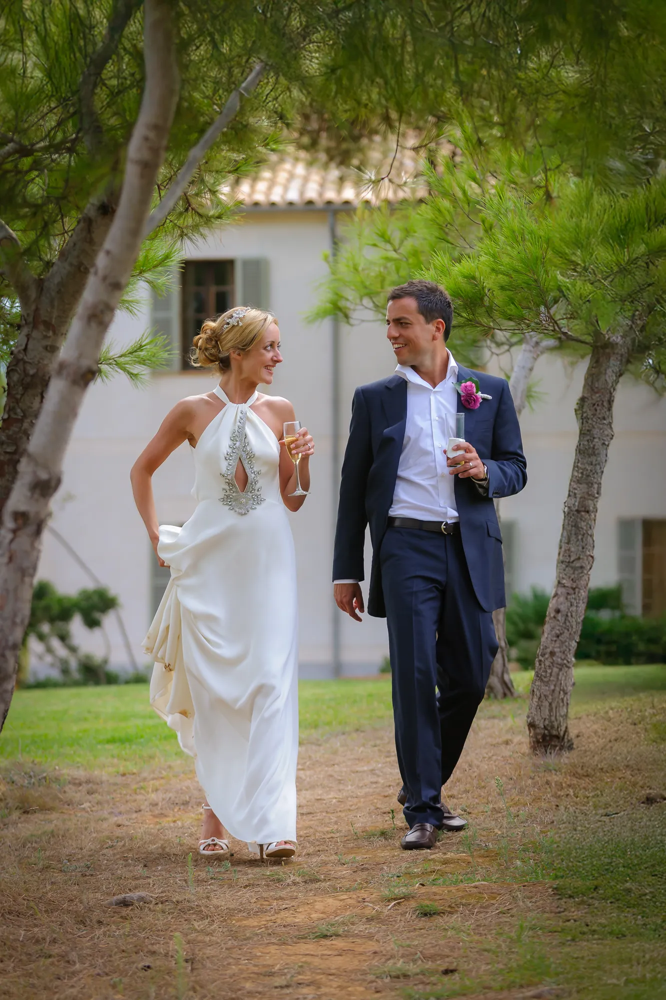 Bride in white dress and groom in dark suit walking outdoors holding drinks and smiling at each other.