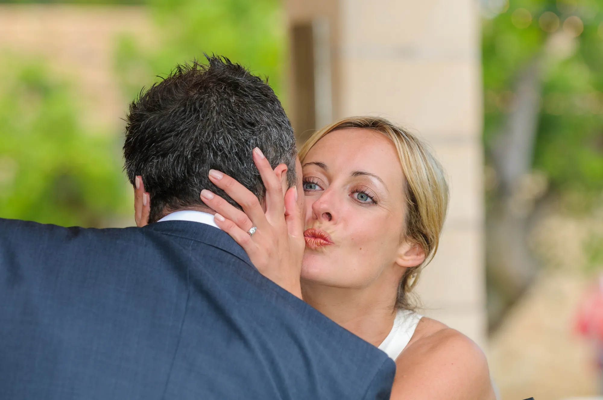 Woman with blonde hair and an engagement ring on her finger embraces and kisses a man in a suit from behind.