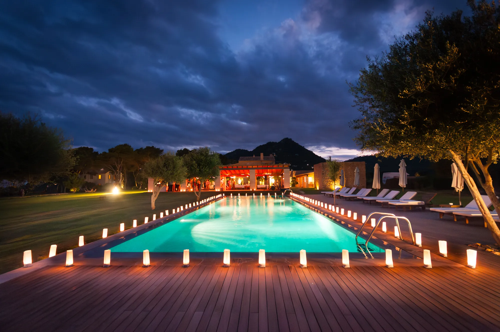 Night view of an illuminated outdoor swimming pool with small lights along the deck, lounge chairs, trees, and a lit pavilion with people in the background.