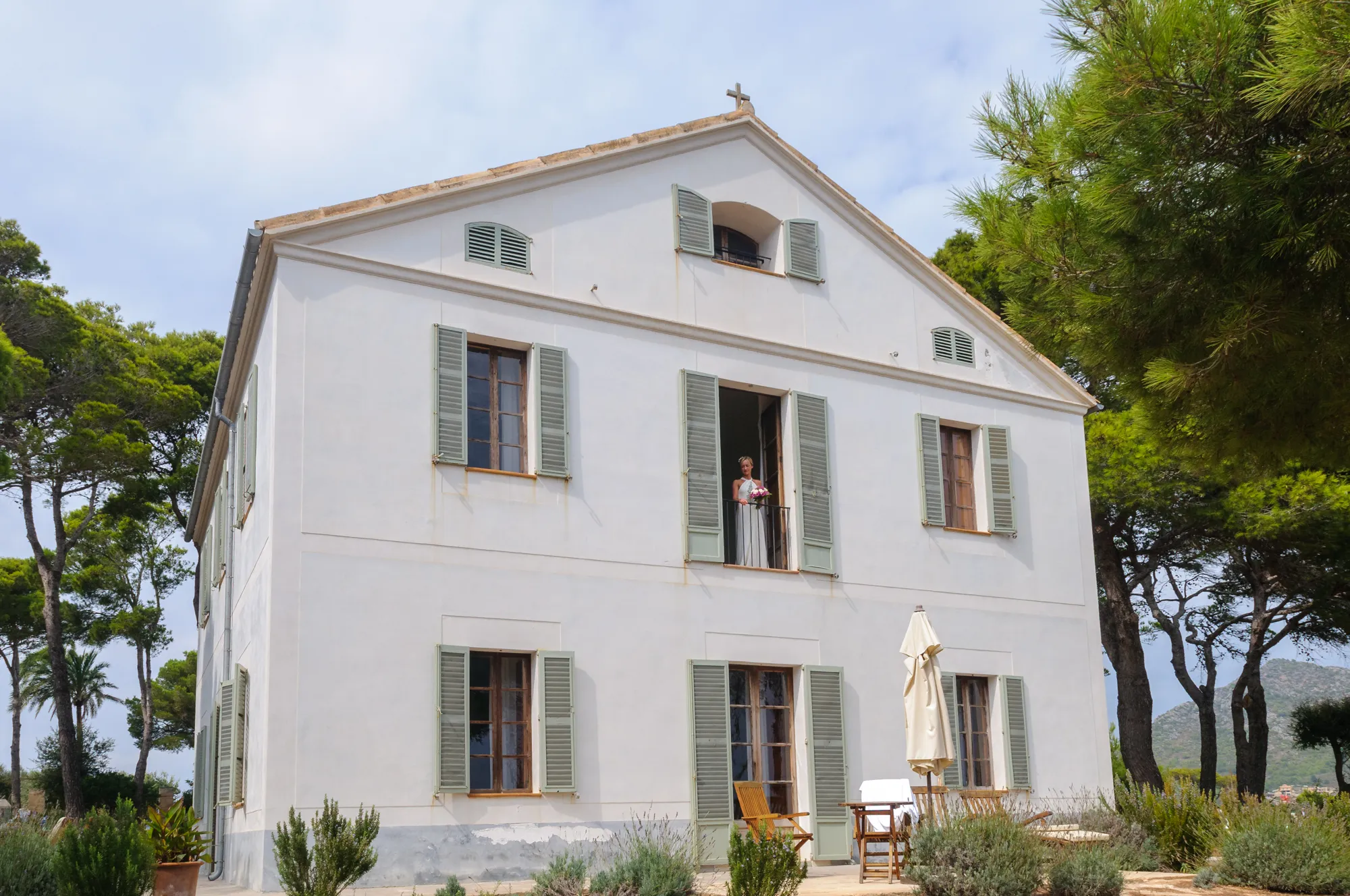 White two-story house with green shutters surrounded by trees, with a woman in a white dress holding flowers standing on a second-floor balcony.