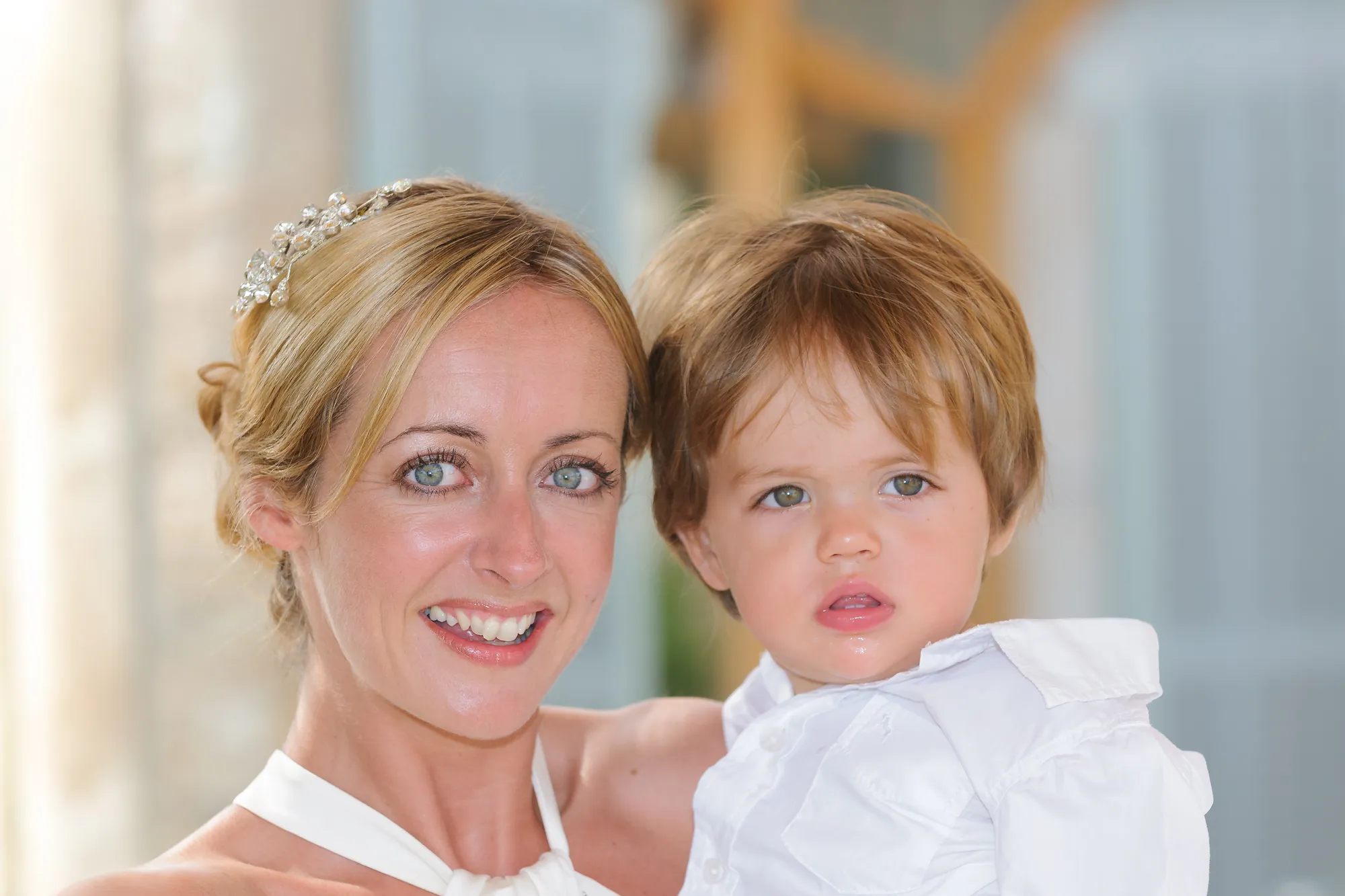 Smiling woman with blonde hair and hair accessory holding a young boy wearing a white shirt.