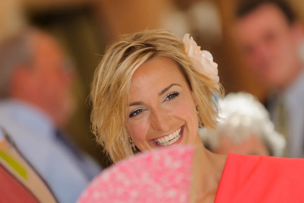 Smiling woman with short blonde hair and a flower hair accessory, holding a pink fan, with blurred people in the background.