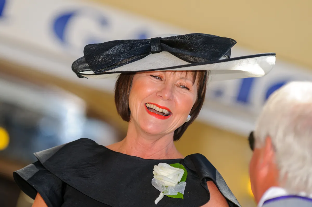 Smiling woman wearing a wide-brimmed black and white hat, black dress, and a white rose boutonniere.