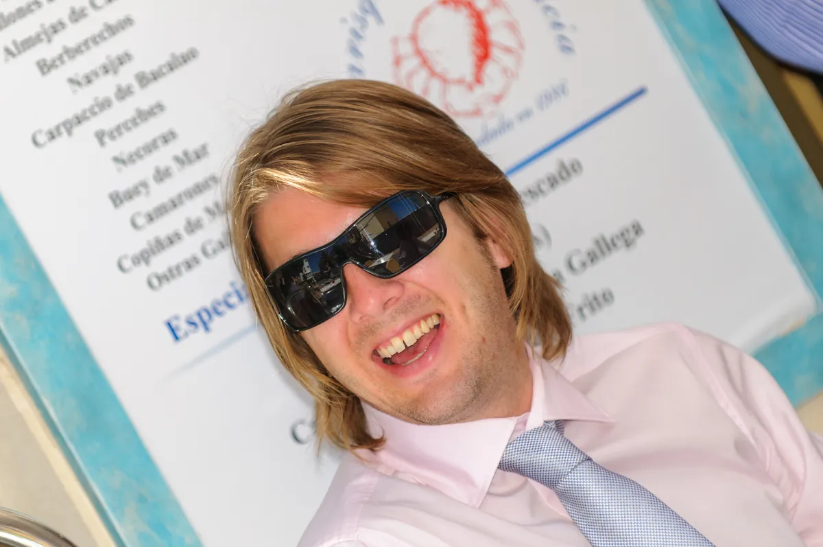 Smiling man with blond hair wearing black sunglasses, a light pink shirt, and a patterned tie in front of a menu board.