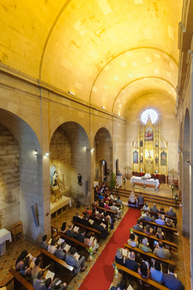 Interior of a church during a wedding ceremony with guests seated on wooden pews and a priest at the altar.