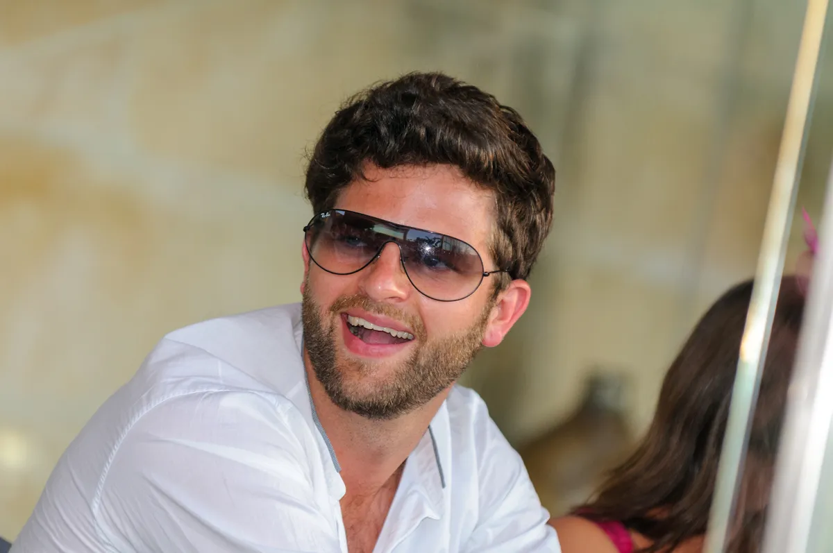 Smiling man with curly brown hair and beard wearing sunglasses and a white shirt.