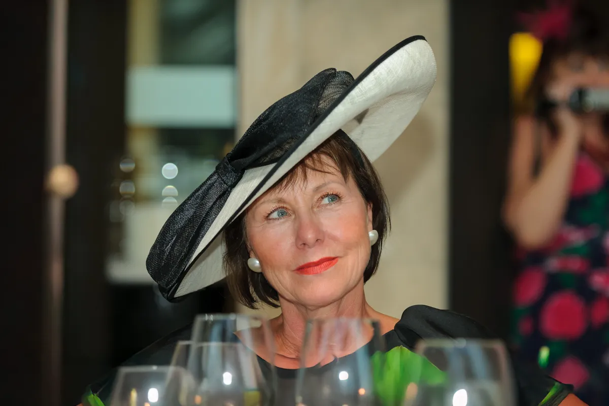 Woman wearing a large black and white hat with pearl earrings, looking upward while seated at a table with glasses.