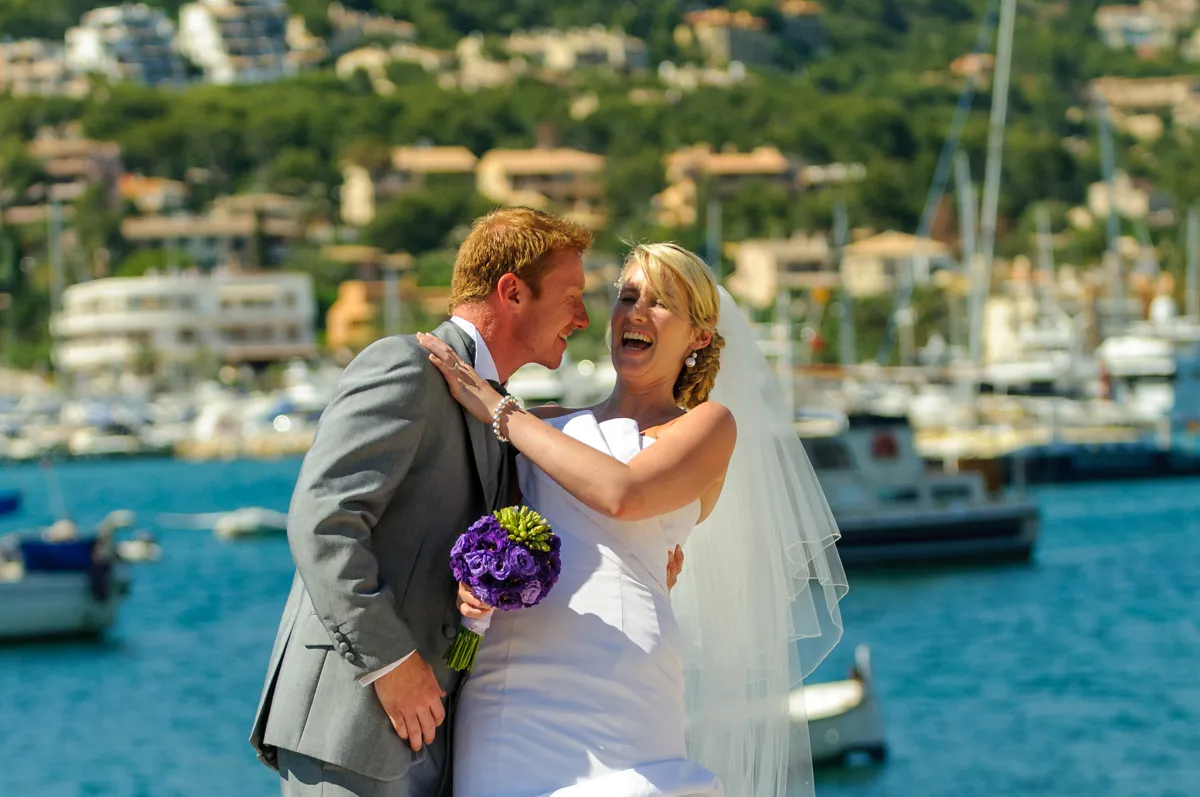 Bride and groom laughing together by the water with boats and hillside buildings in the background.