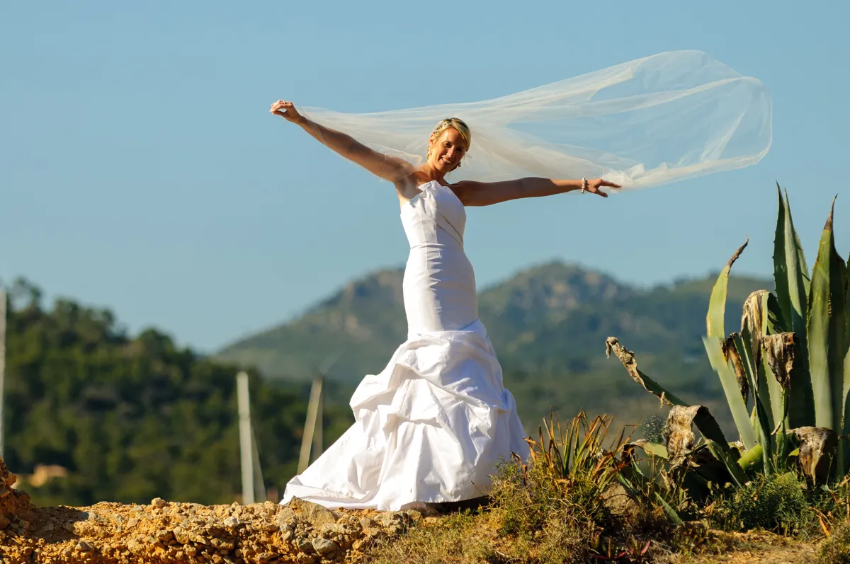 Bride in a white strapless wedding dress stands outdoors with arms outstretched holding a flowing veil against a mountainous background.