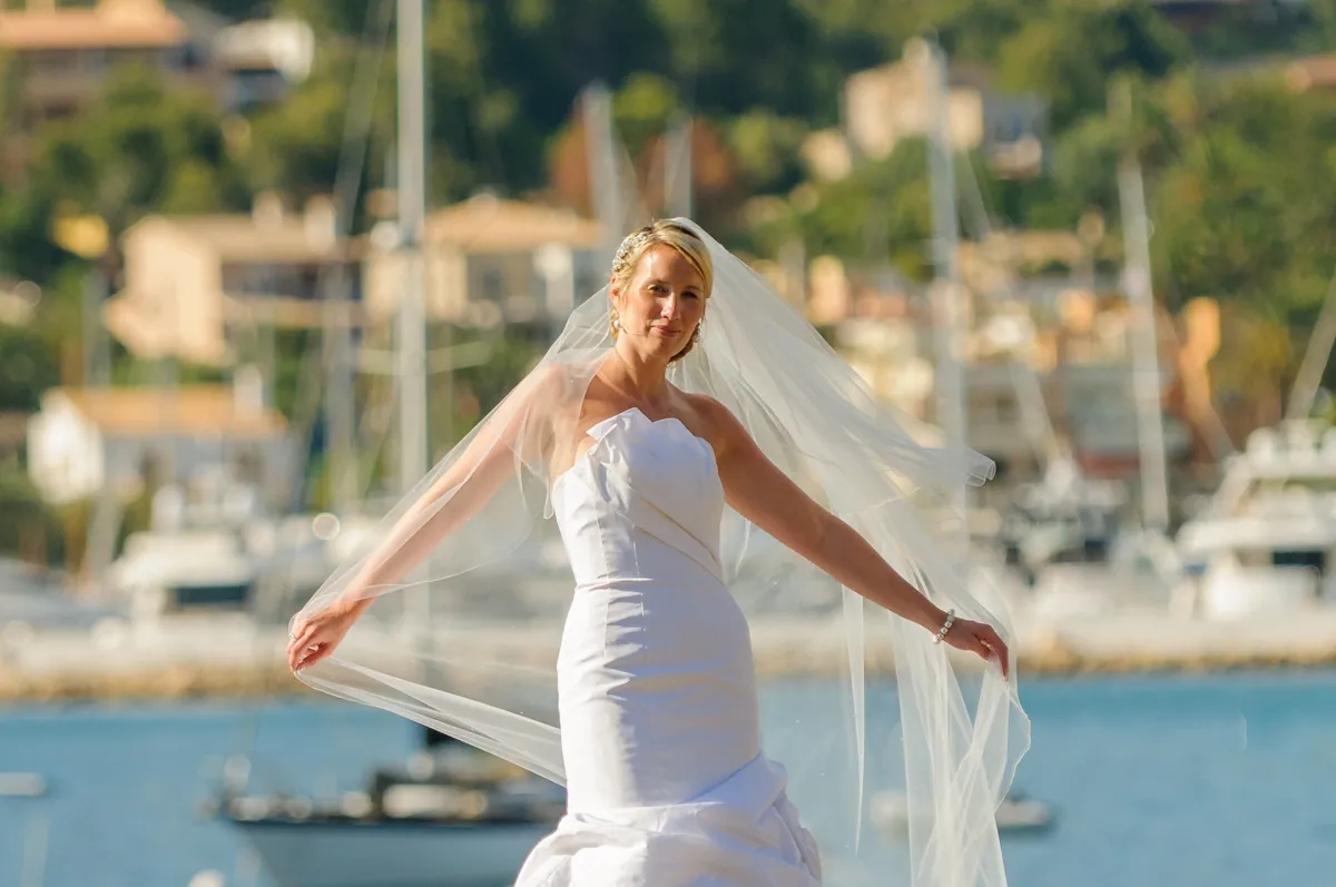 Bride in a strapless white wedding dress holding out her veil with boats and waterfront buildings in the background.