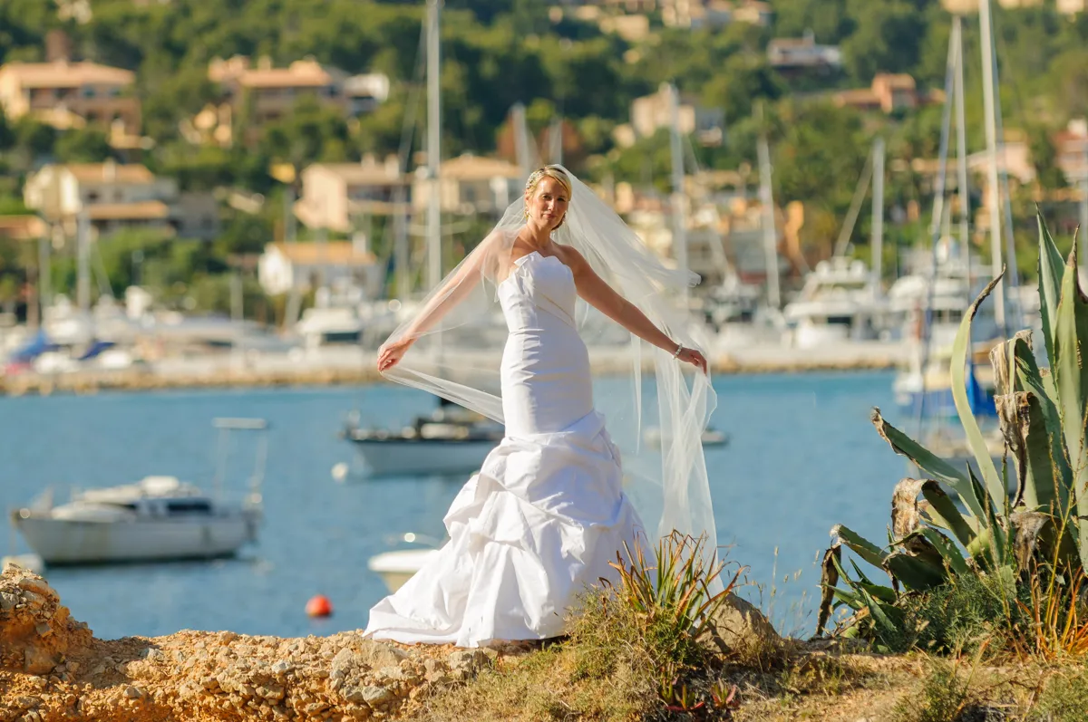 Bride in a white wedding gown holding her veil by the seaside with boats and buildings in the background.