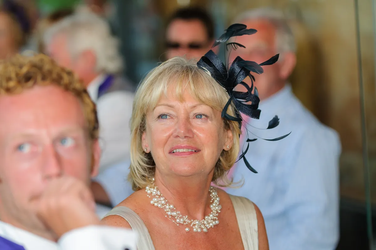 Smiling woman wearing a black fascinator and pearl necklace at a social event with blurred people in the background.