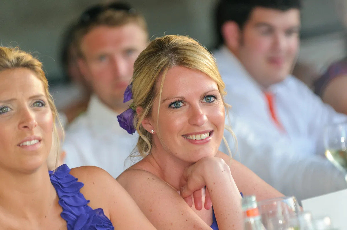 Smiling blonde woman with purple flowers in her hair wearing a purple dress, sitting at a table with glasses and people blurred in the background.