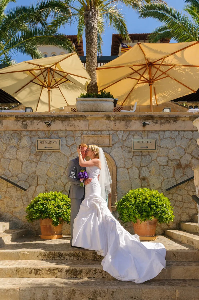Bride in white gown kissing groom in gray suit on stone steps with large umbrellas and palm trees in background.