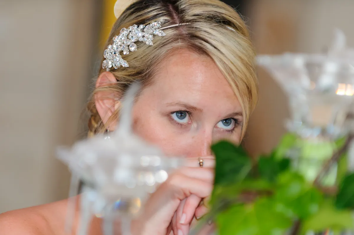 Close-up of a woman with blonde hair and a jeweled hair accessory, looking intently over green foliage and glass candle holders.