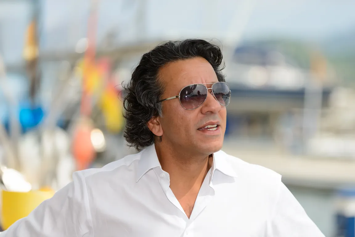 Man wearing sunglasses and a white shirt speaking outdoors near a marina.