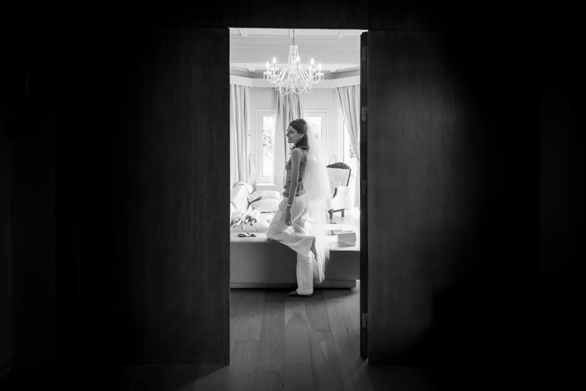 Black and white photo of a smiling bride adjusting her shoe in a bright living room with a chandelier and curtains, seen through partially open dark doors.