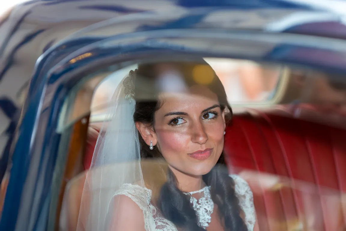 Bride with veil and lace dress sitting in a car with red leather seats, viewed through the window.