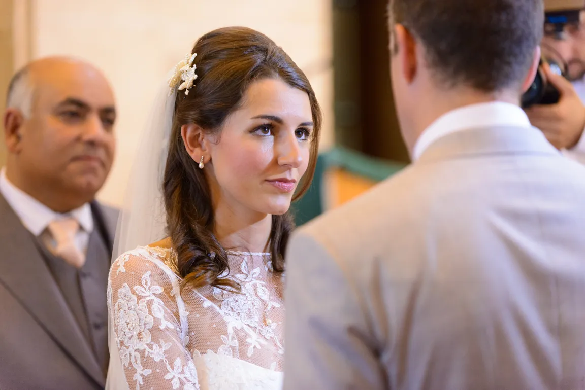 Bride in lace wedding dress looking at groom during wedding ceremony with an older man in background.