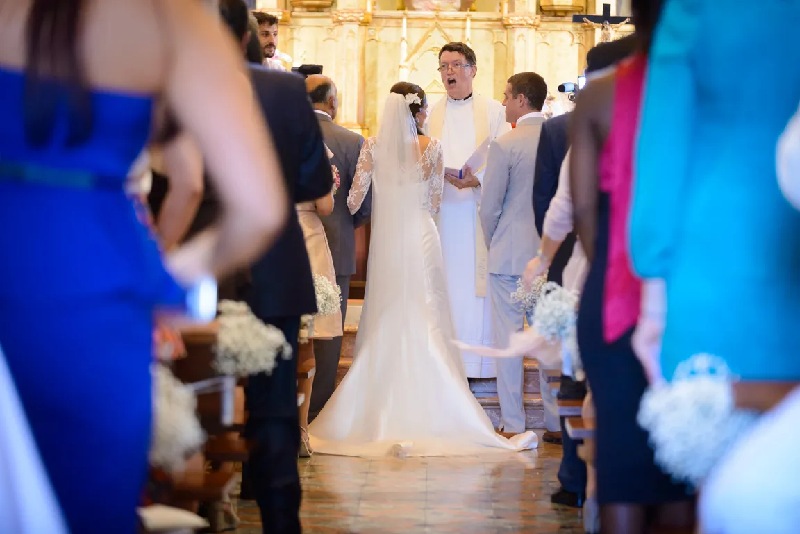 Bride and groom standing before a priest in a church during a wedding ceremony with guests seated on both sides.