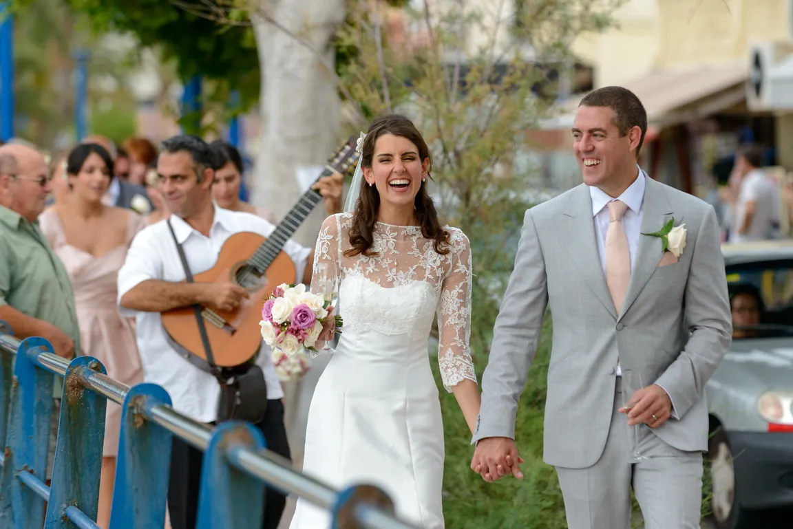 Bride and groom holding hands and smiling, with bride holding a bouquet and a man playing guitar behind them.