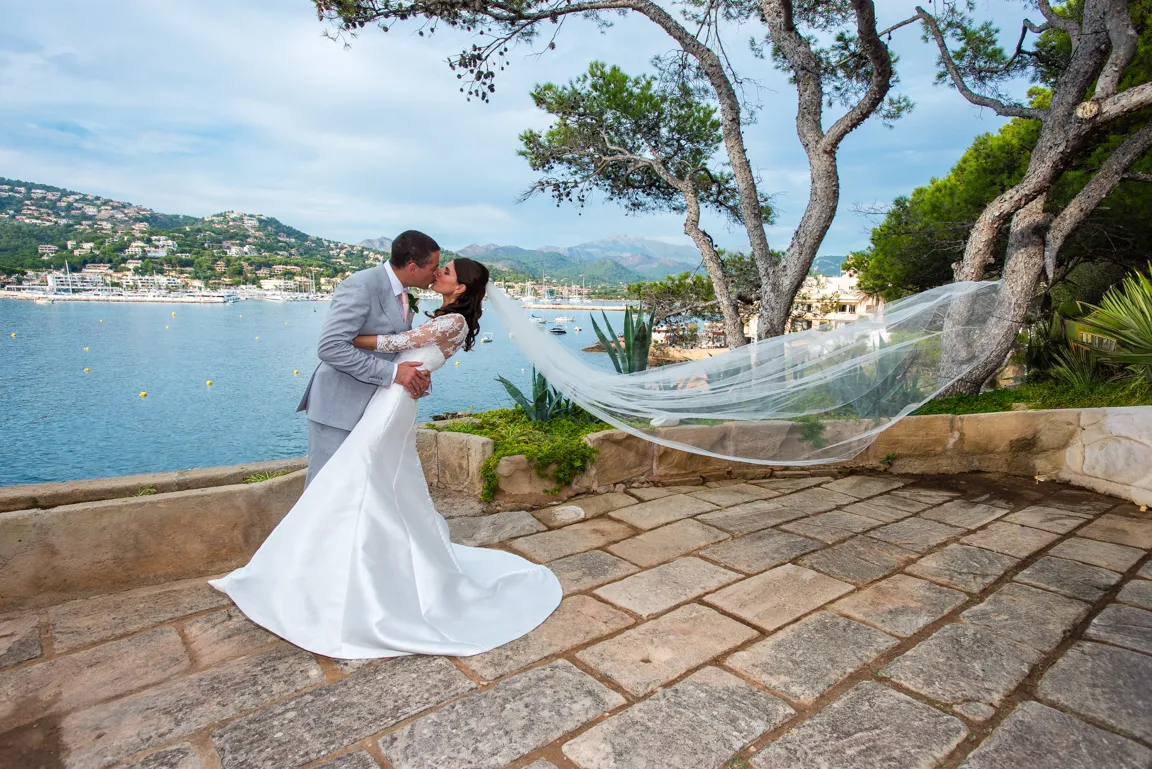 Bride and groom kissing on a stone terrace by the sea with the bride’s veil flowing behind her.