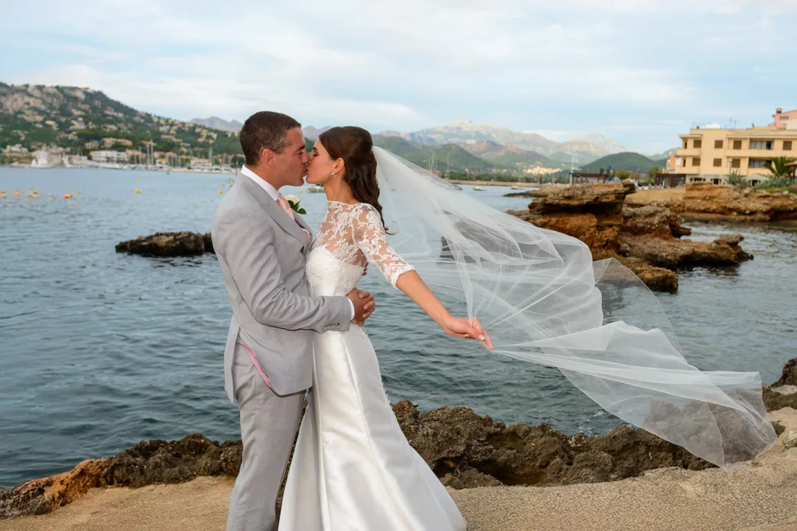 Bride and groom kissing by the rocky shore with mountains and buildings in the background, bride's veil flowing in the wind.