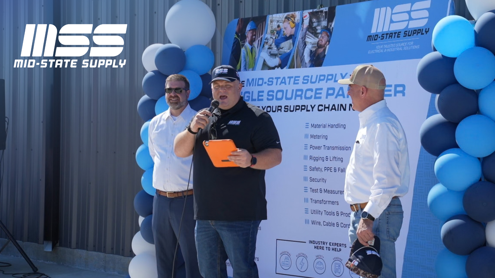 Man in a black shirt holds microphone in front of blue balloons at the Mid-State Supply Geismar Ribbon Cutting
