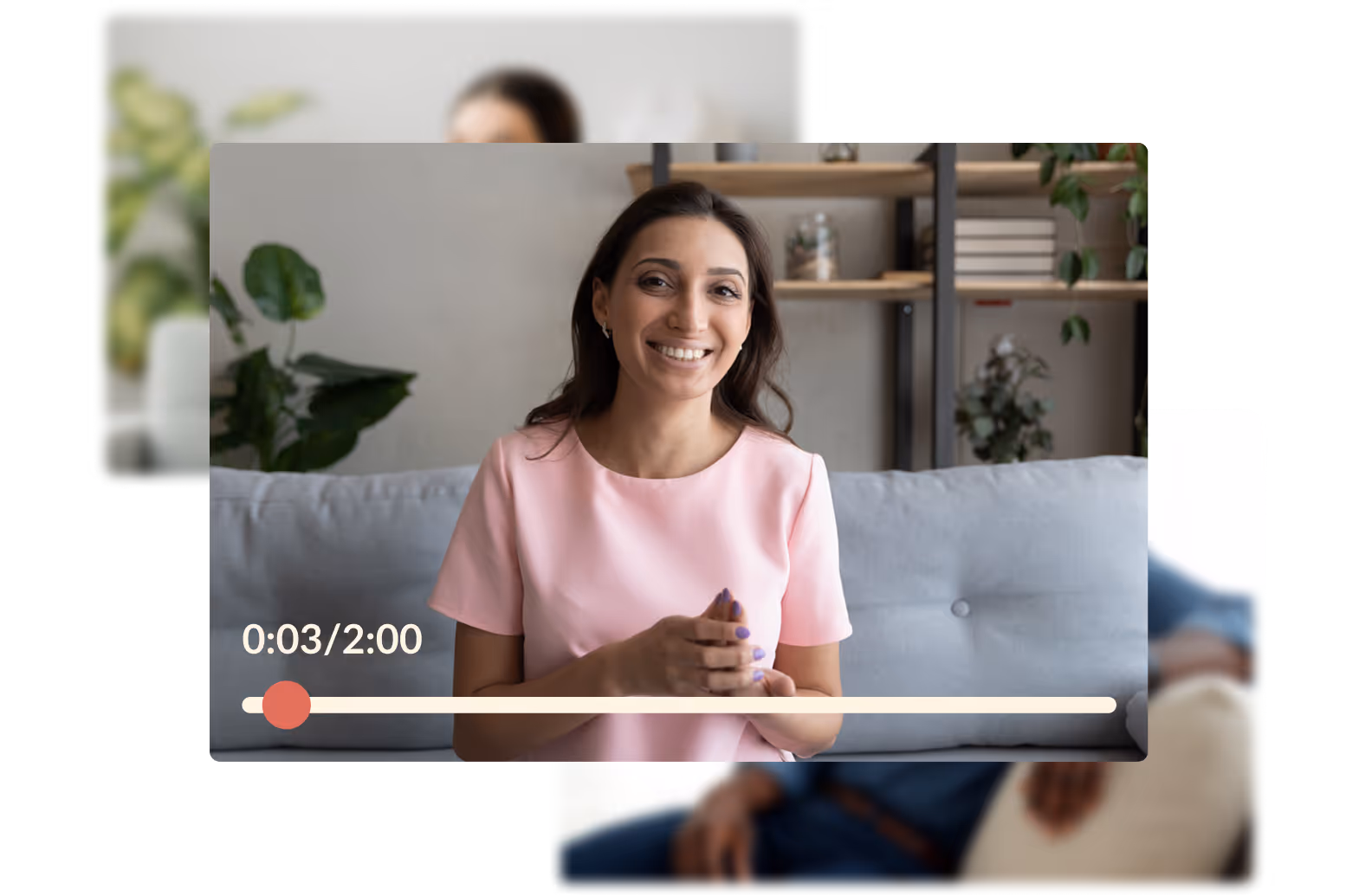 Smiling woman in pink shirt sitting on a gray couch with plants and shelves in the background, video progress bar at 0:03 of 2:00.