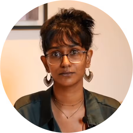 Person with glasses, dark hair in a bun, wearing heart-shaped earrings and a dark shirt in an indoor setting.