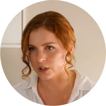 Woman with light skin and red hair styled in loose curls, wearing a white shirt and speaking.