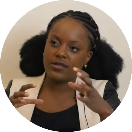 Woman with braided hair and a light blazer speaking and gesturing with her hands.