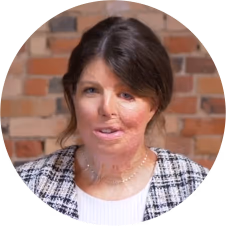 Woman with dark hair styled back, wearing a white top and checkered jacket, against a brick wall background.