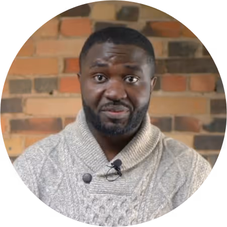 Man with short hair and beard wearing a light gray cable-knit sweater in front of a brick wall.
