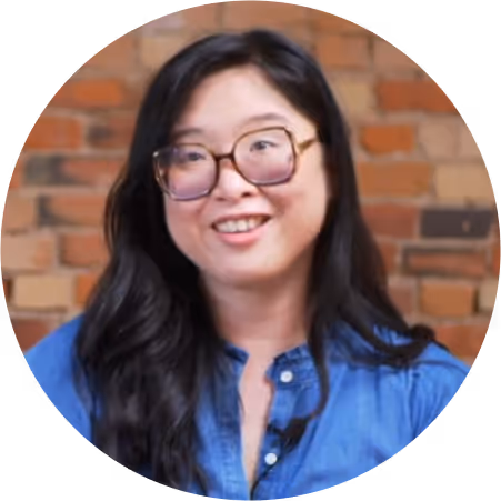 Smiling woman with long black hair and large glasses wearing a blue shirt against a brick wall background.