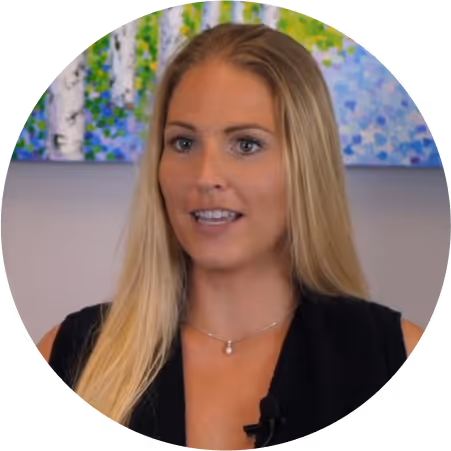 Blonde woman with long hair wearing a black top and pendant necklace speaking in front of a colorful painting.