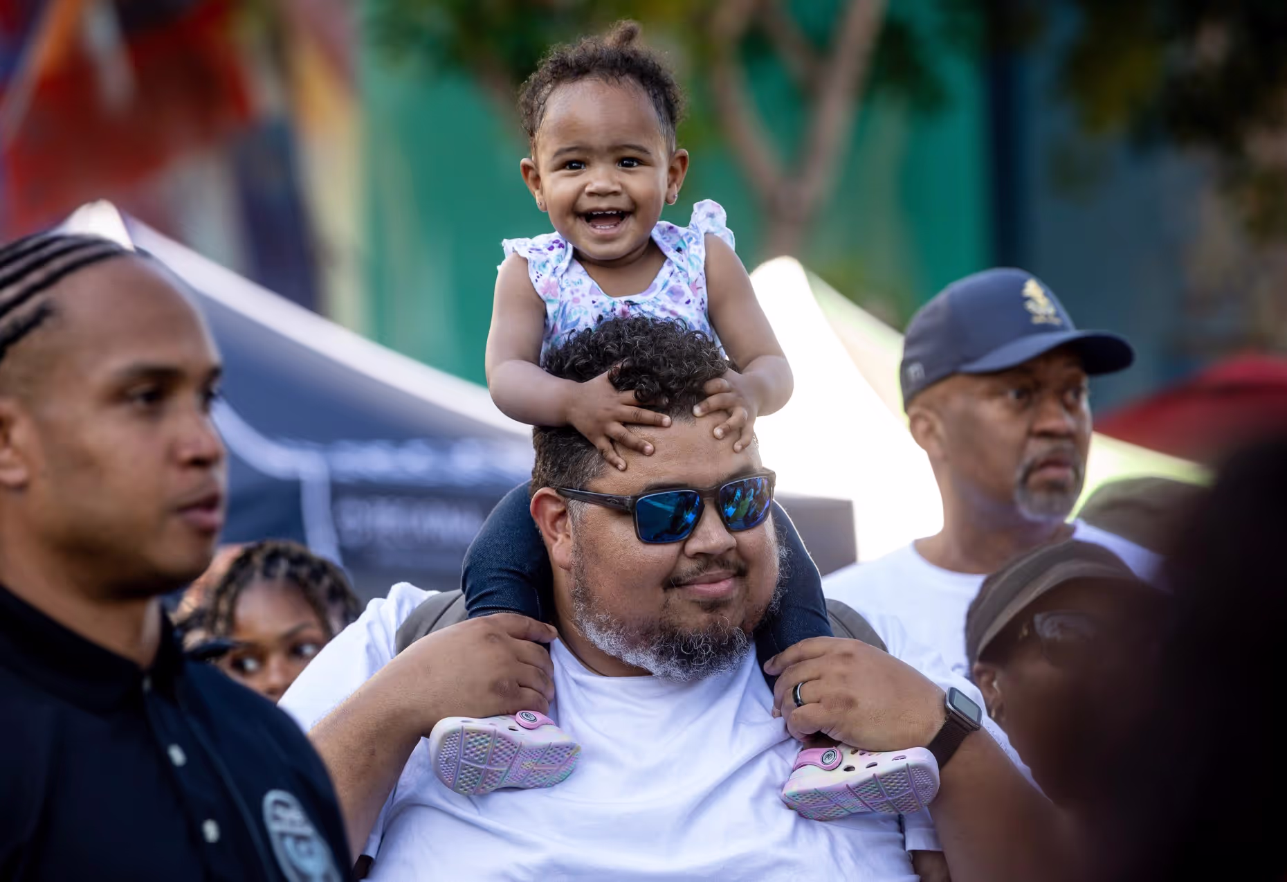 A smiling toddler sits on a man's shoulders in the festival crowd.