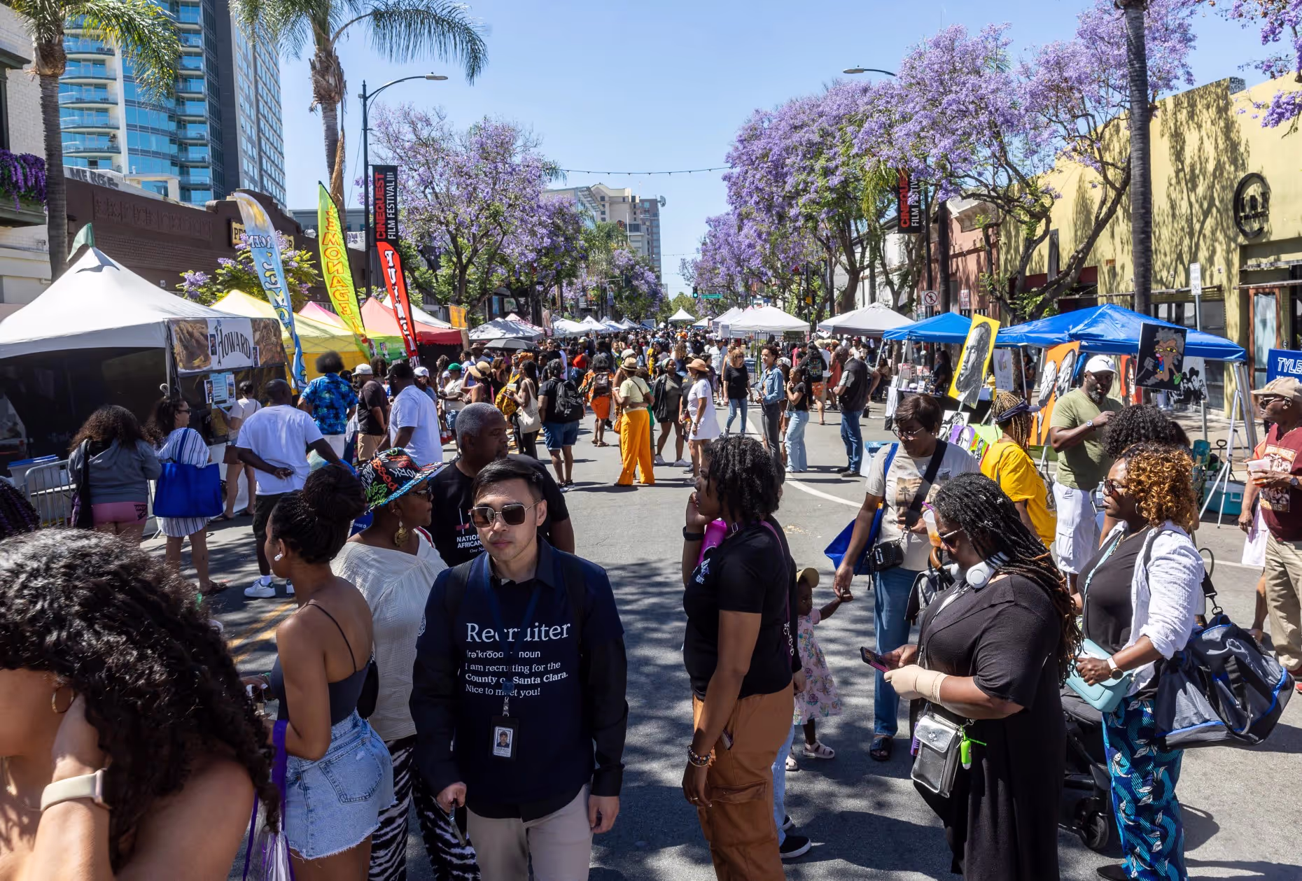 Festival crowds walk between vendor tents on a street lined with purple jacaranda trees.