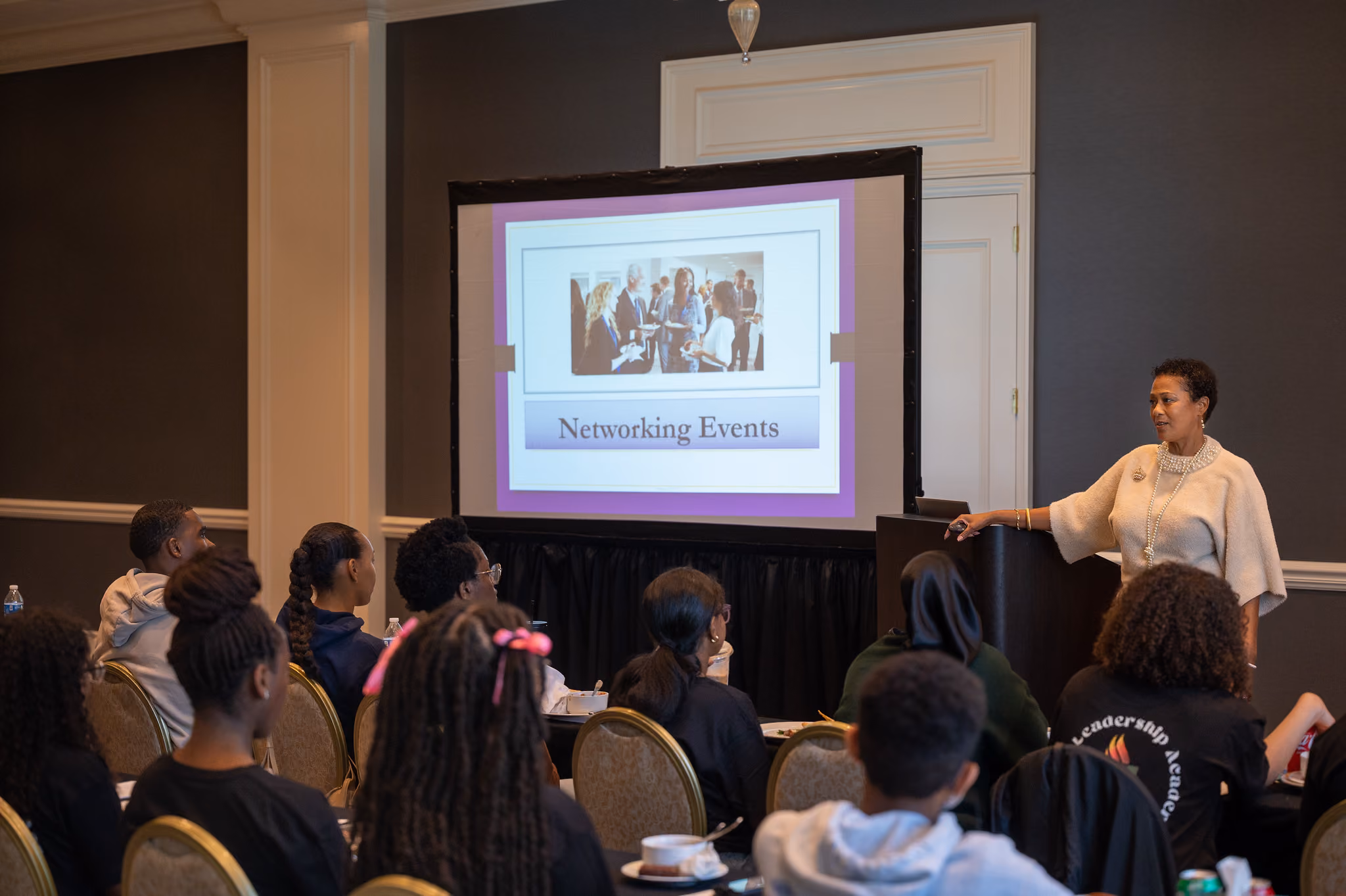 A presenter speaks beside a projection screen reading "Networking Events" while students sit at tables.
