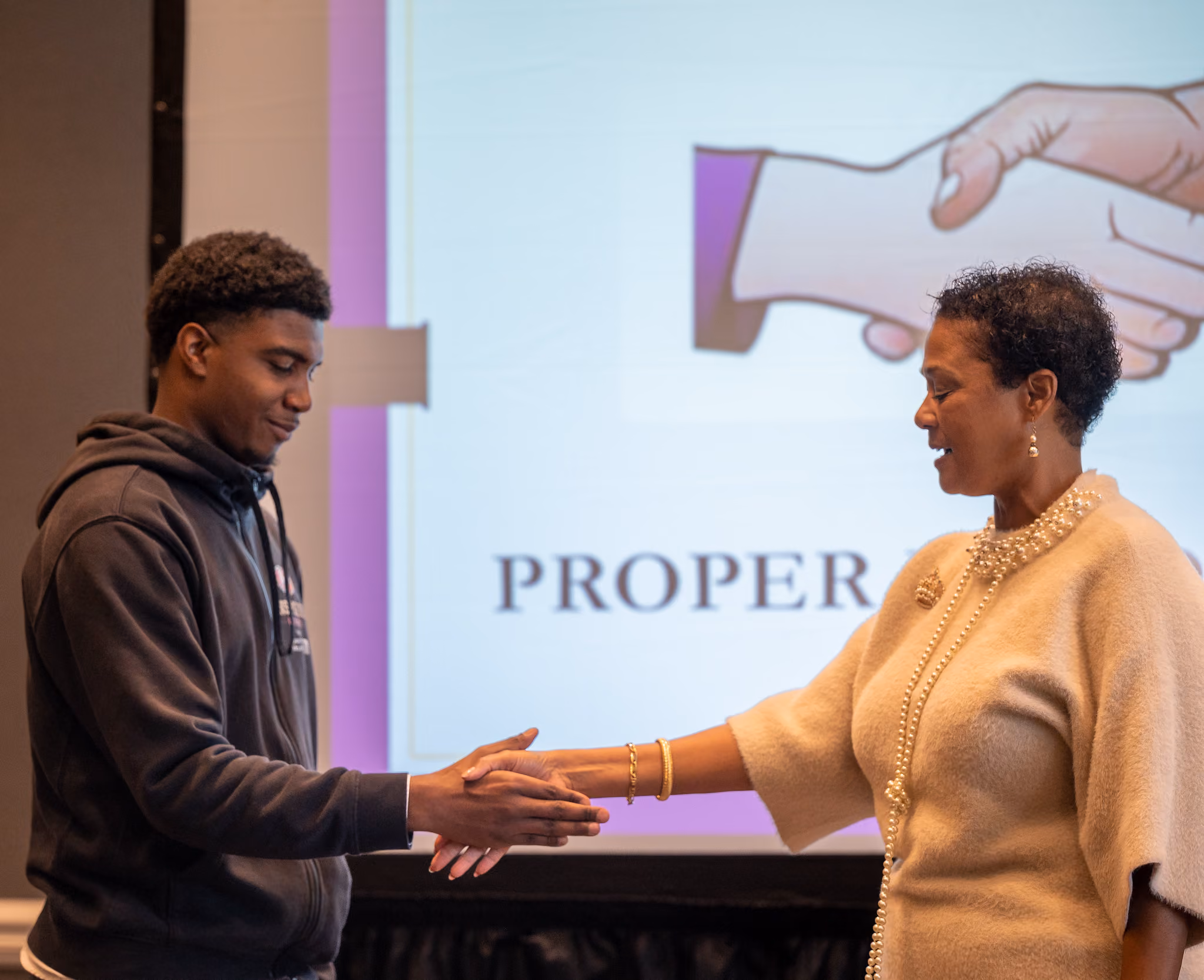 A young man in a dark hoodie shakes hands with a woman in a cream sweater in front of a projection screen.