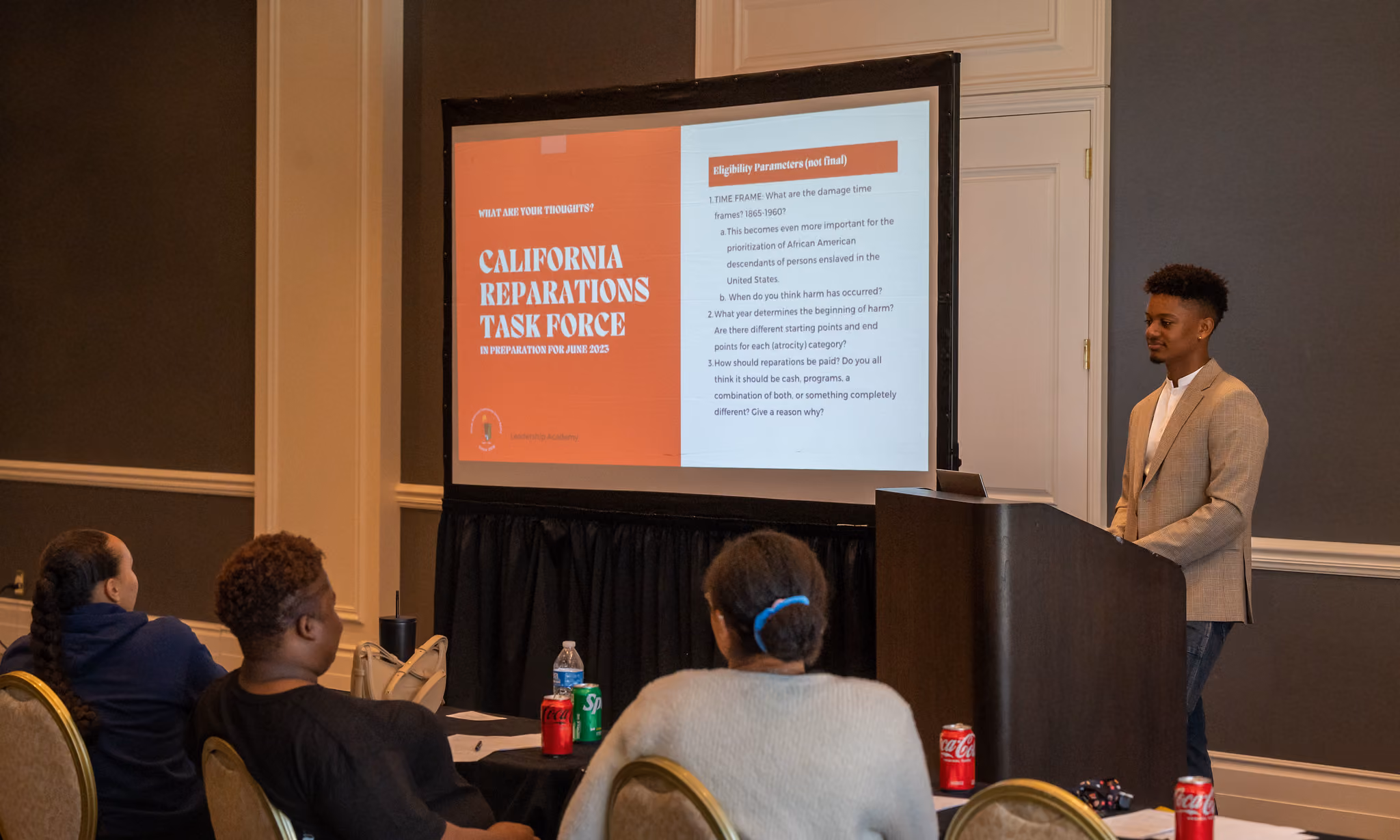 A young presenter stands at a podium beside a slide titled "California Reparations Task Force" while students watch.