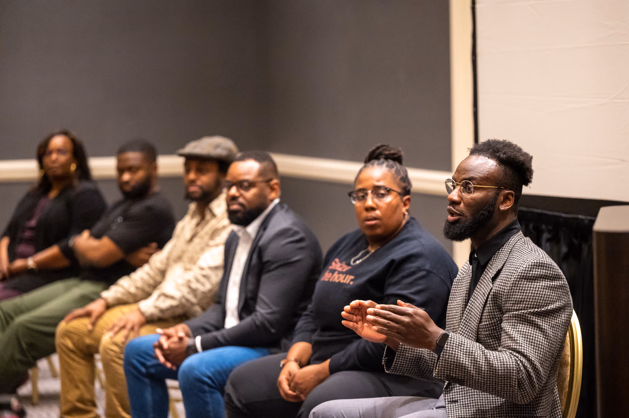 Panelists seated in a row listen as a bearded man in a houndstooth blazer speaks.