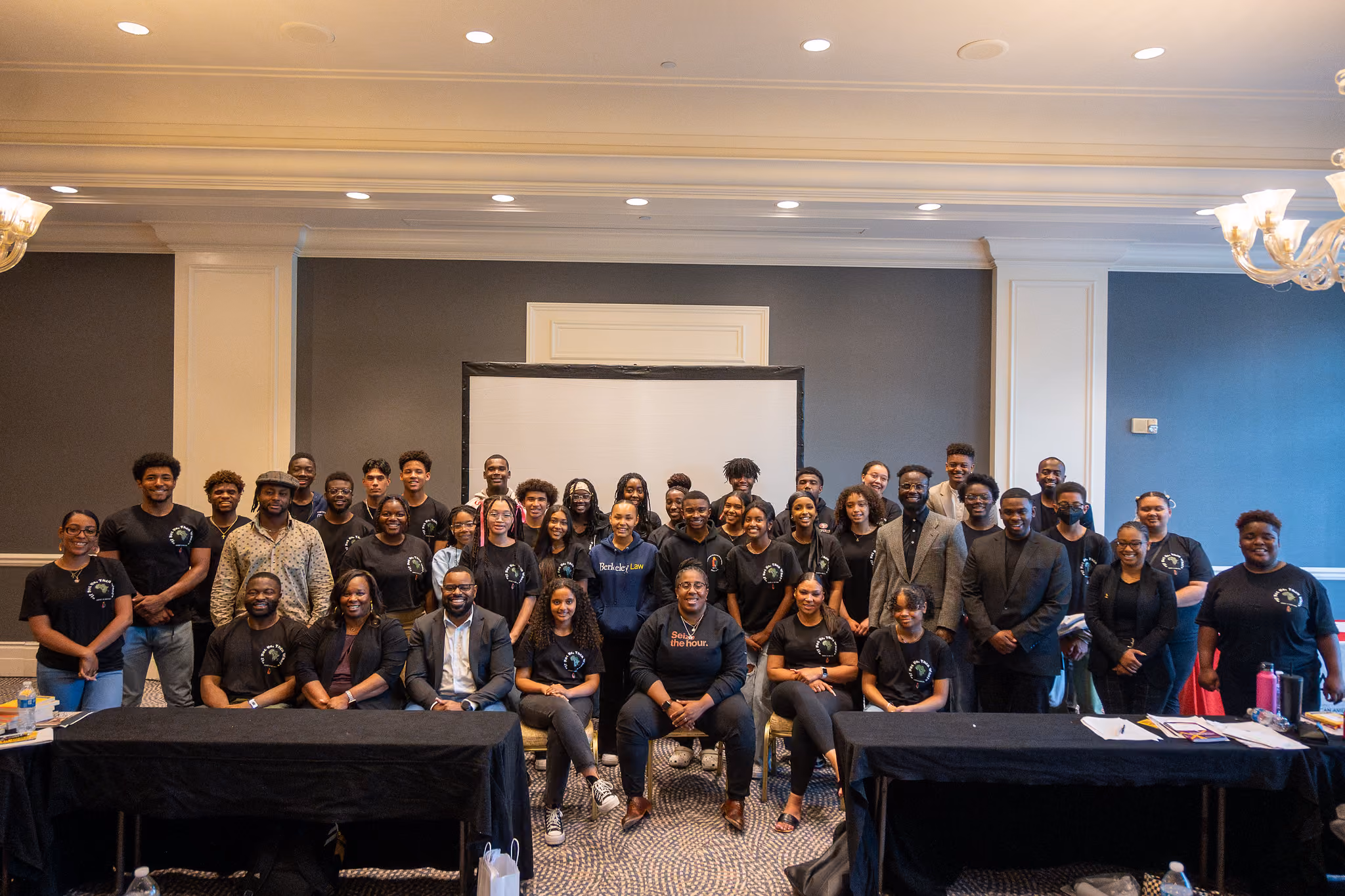 A large group of participants in black shirts poses together in a ballroom.